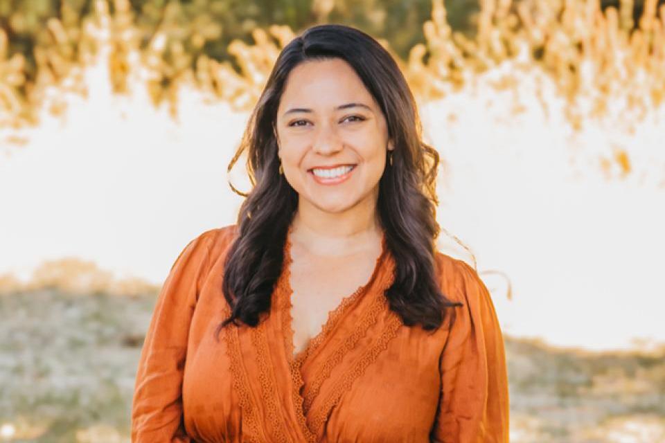 A woman with long dark hair, wearing an orange blouse, stands outdoors smiling, with blurred greenery and sunlight in the background.
