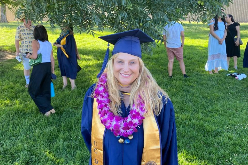 A graduate in a cap and gown with a purple flower lei stands on grass under a tree; other people are gathered in the background at an outdoor event.