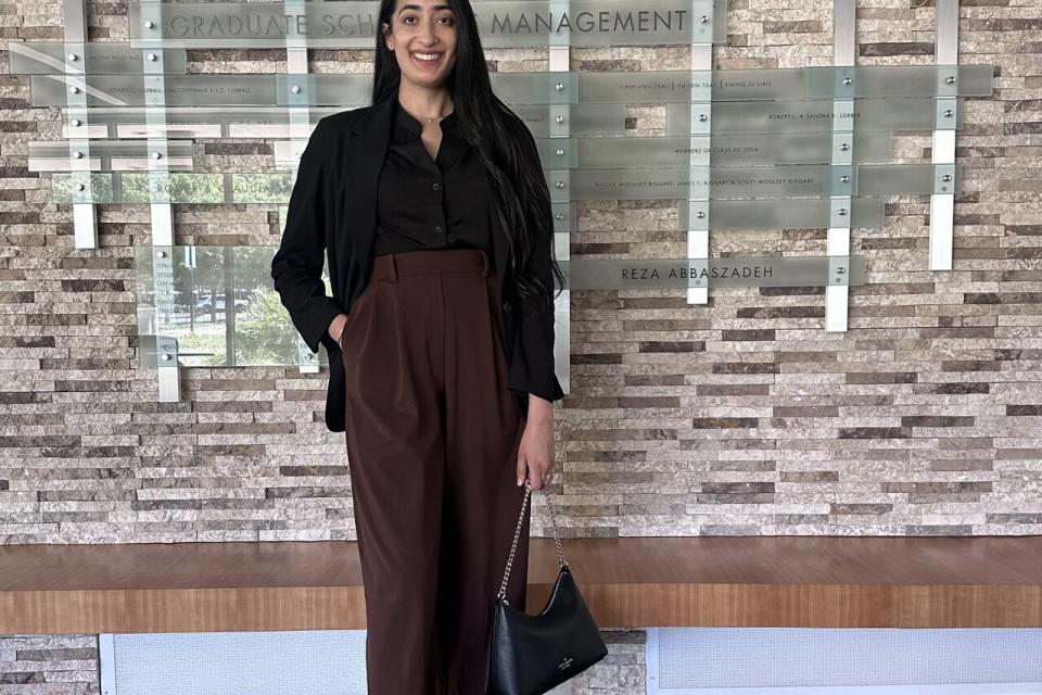A woman stands indoors in business attire, holding a handbag and smiling, in front of a wall displaying names and a sign reading “Graduate School of Management.”