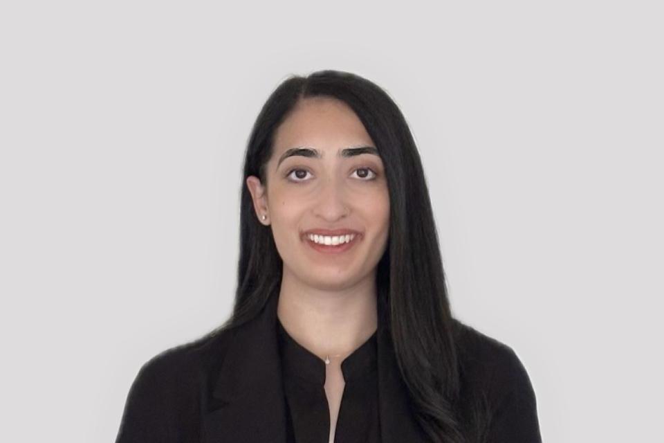 A woman with long dark hair, wearing a black blazer and dark shirt, stands in front of a plain light gray background, facing the camera and smiling.