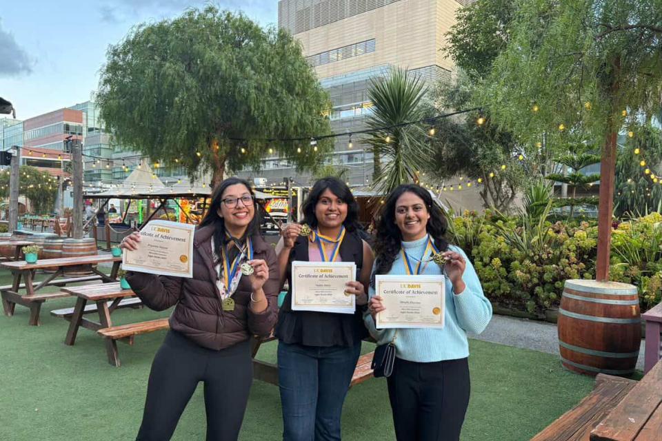 Three women stand outdoors holding certificates and wearing medals, smiling at the camera. There are picnic tables, plants, and a building in the background.