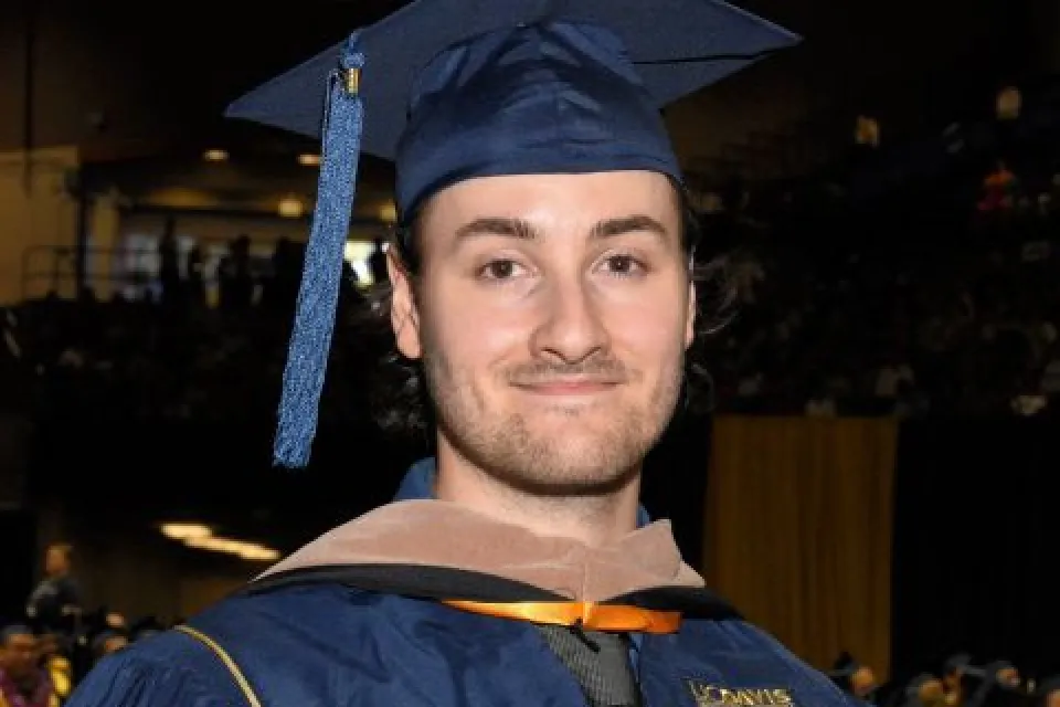 A person in a blue graduation cap and gown stands indoors at a graduation ceremony, with other graduates and an audience visible in the background.