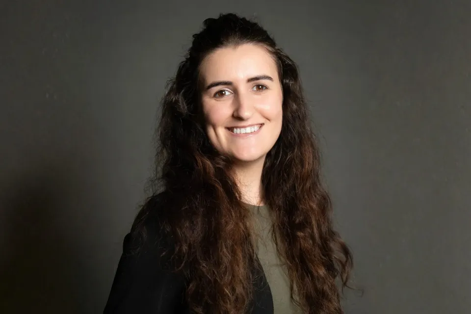 A woman with long dark hair smiles at the camera against a grey background.