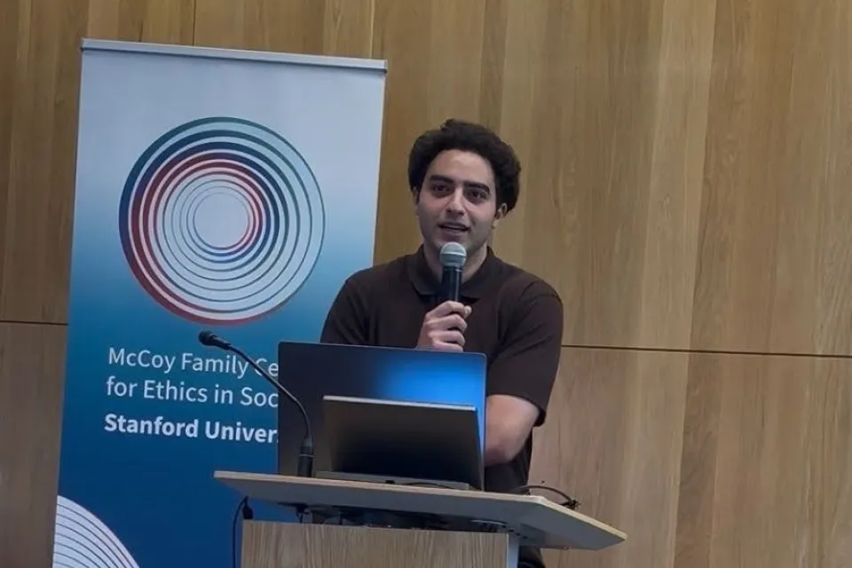 A man speaks into a microphone at a podium in front of a Stanford McCoy Family Center for Ethics in Society banner.